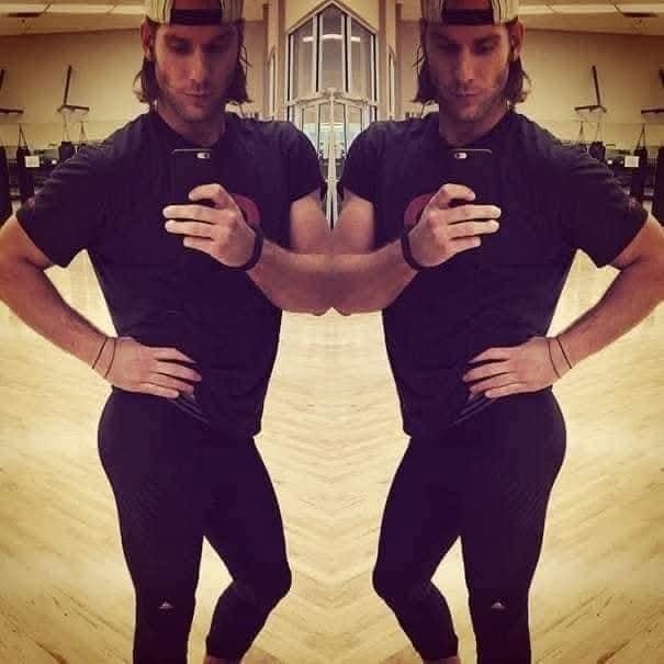 Osoba v posilňovni pred zrkadlom, oblečená v športovom tričku a nohaviciach, fotografuje svoj odraz.