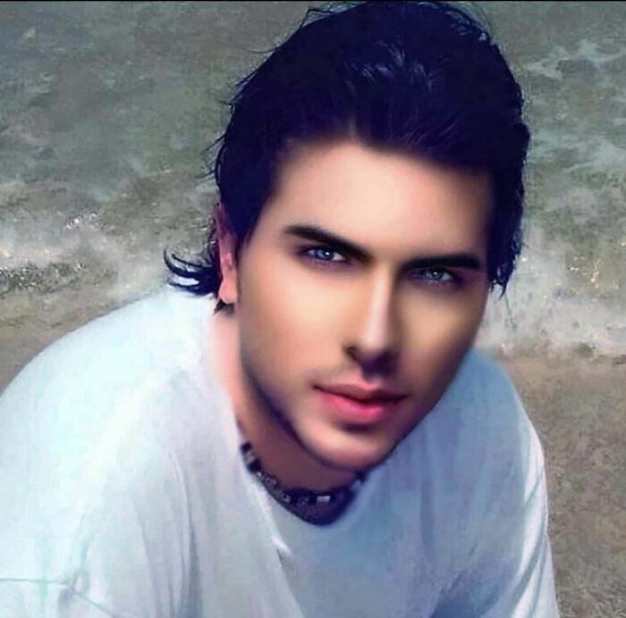 Ľudia pri mori, mladý muž s tmavými vlasmi a bielym tričkom na pláži, pozadie s pieskom a vodou.