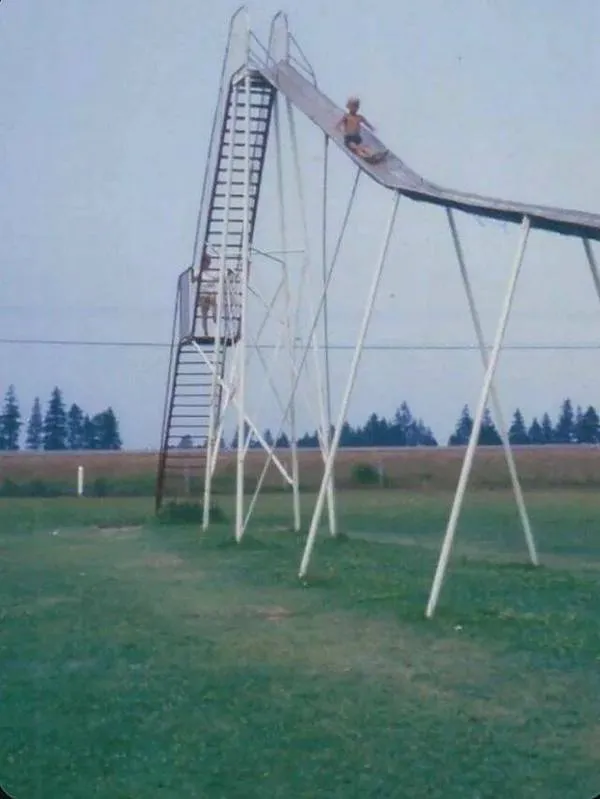 Dlhá šmykľavka v parku, dieťa sa šmýka dolu; letný deň, trávnatý priestor.