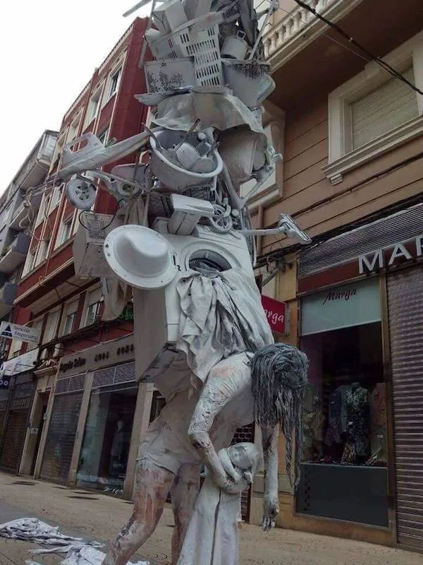 Socha z domácich spotrebičov a postavy v bielom v meste medzi budovami.