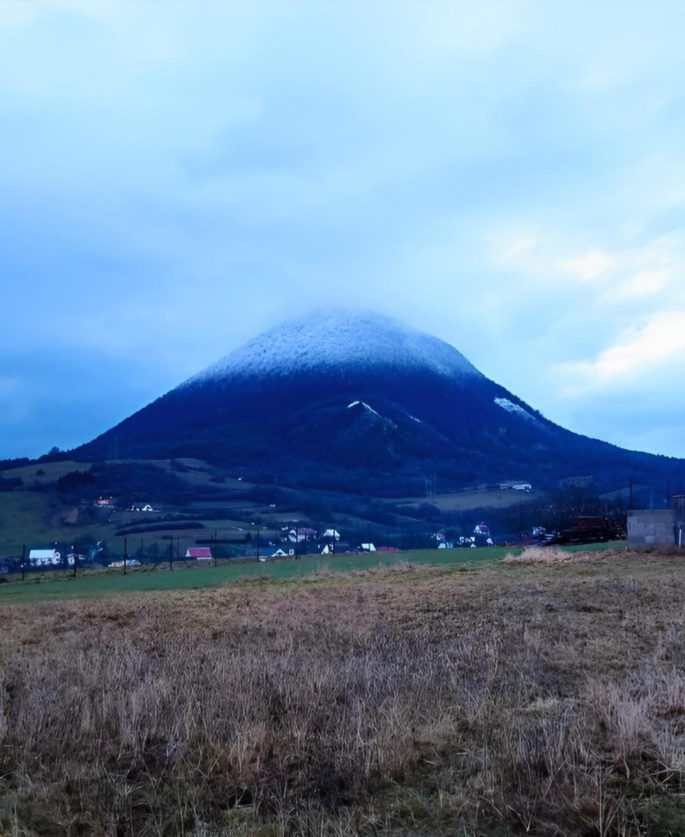 Hora snehom pokrytá na vrchole, obklopená dedinou a poľami pod zamračenou oblohou v zimnom podvečeri.
