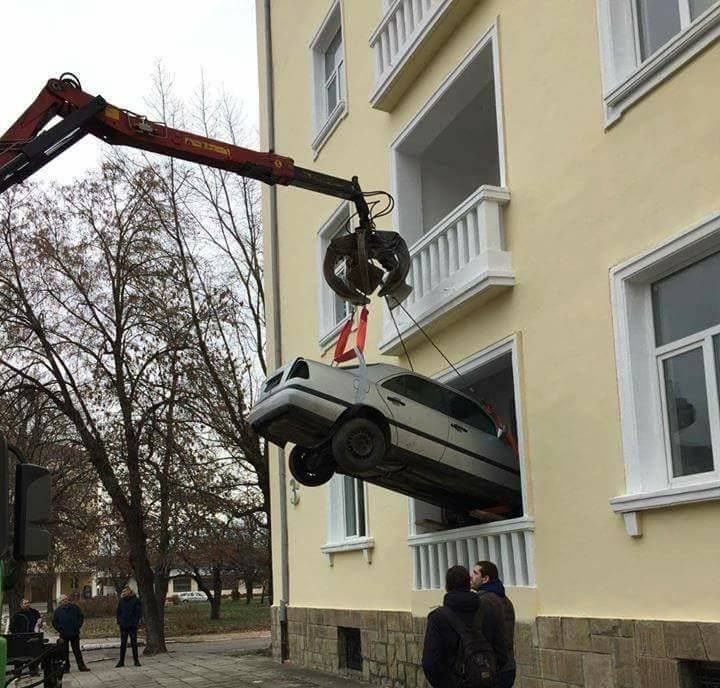 Auto je žeriavom vyťahované cez okno budovy, pozorujú to okoloidúci.