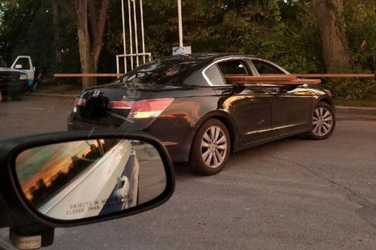 Čierne auto s drevenými doskami trčiacimi z okien na parkovisku, pohľad z bočného zrkadla iného vozidla.