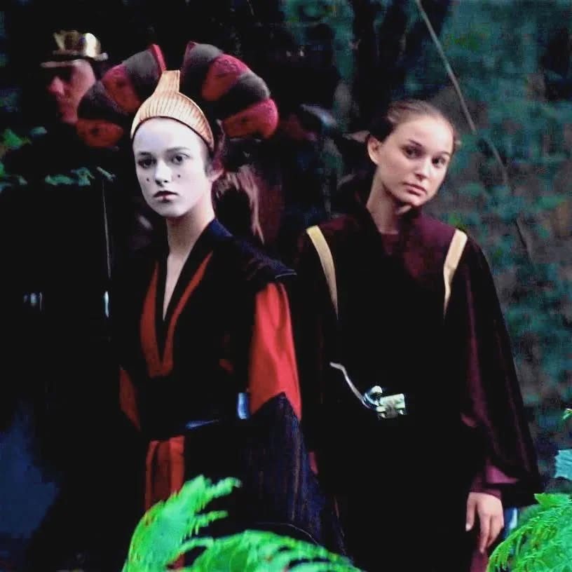 Dve ženy v tradičných šatách stoja v lese, jedna s nalíčenou tvárou a ozdobným účesom, druhá s hladkým účesom.