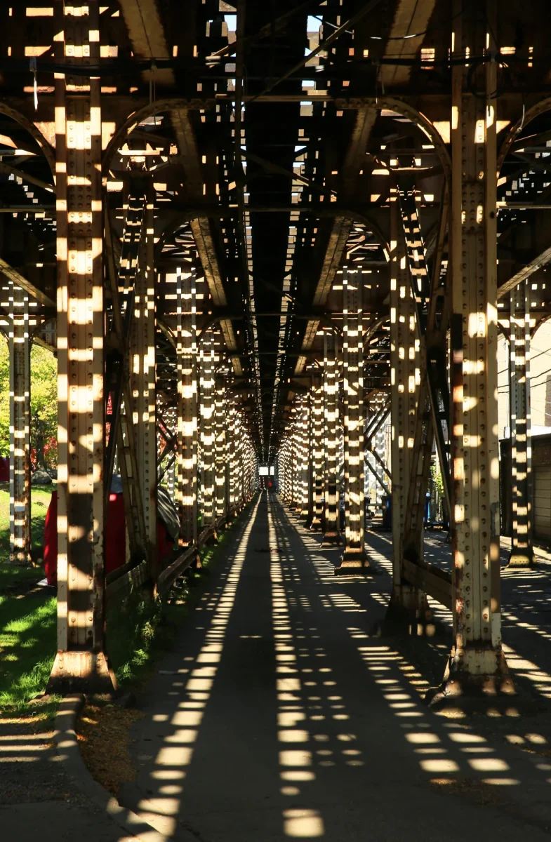 Pohľad na oceľové podpery železničného mosta s tieňmi premietanými cez slnečné svetlo na chodník.