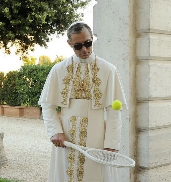 Muž v bielom rúchu hrá na záhrade so slnečnými okuliarmi a tenisovou raketou, s loptičkou vo vzduchu.