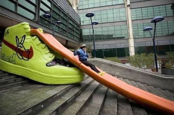 Muž na oranžovej šmykľavke vo forme obrovskej tenisky na mestských schodoch.