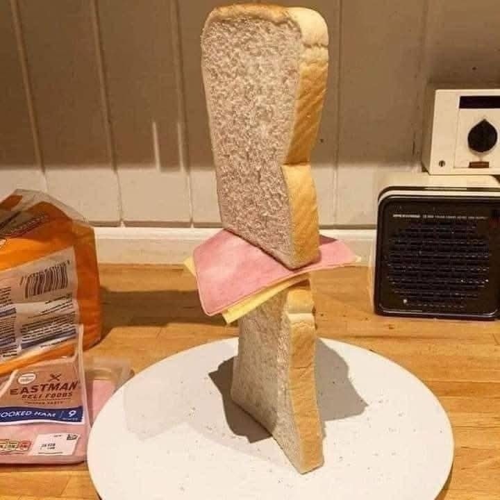 Nezvyčajná sendvičová veža postavená na tanieri z chleba, syra a šunky v kuchynskom prostredí.