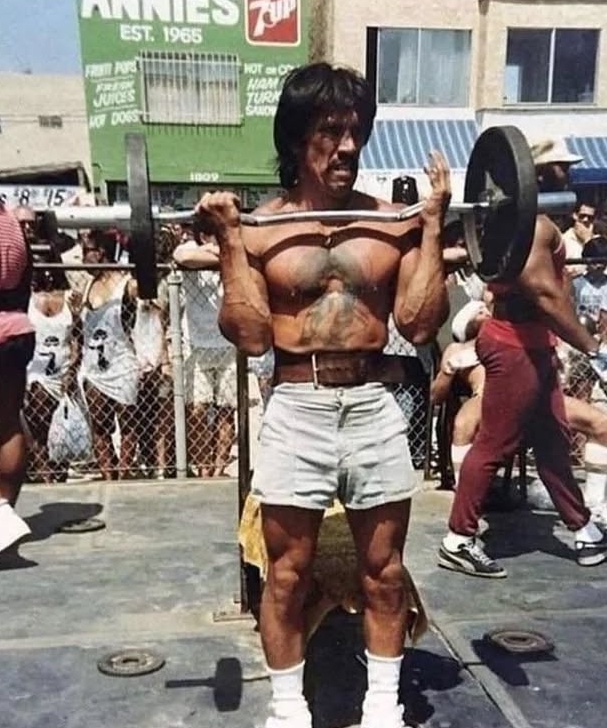 Muž vzpiera činku na vonkajšej posilňovni, oblečený v džínových šortkách, obklopený davom divákov.