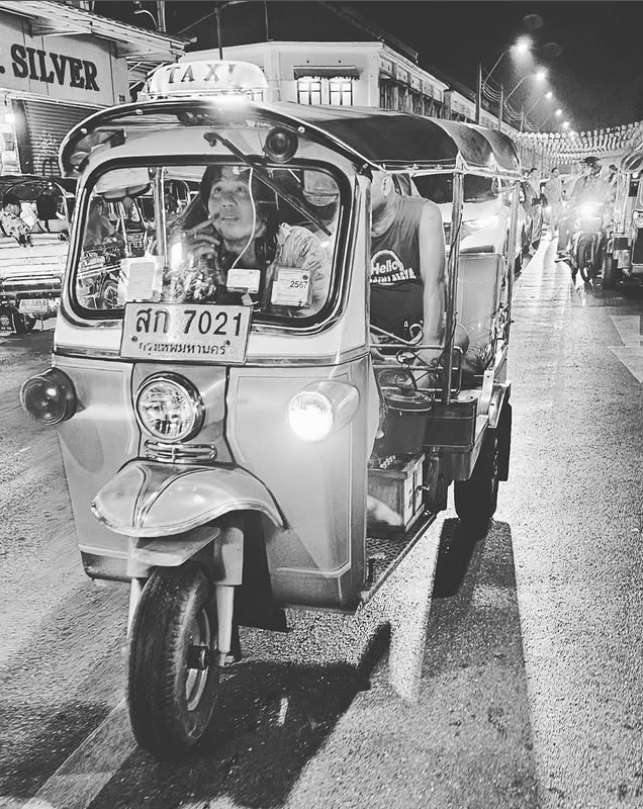 Tuk-tuk v ruchu nočného mesta, vodič vpredu, cestujúci sediaci vzadu. Ulica plná svetiel a života.