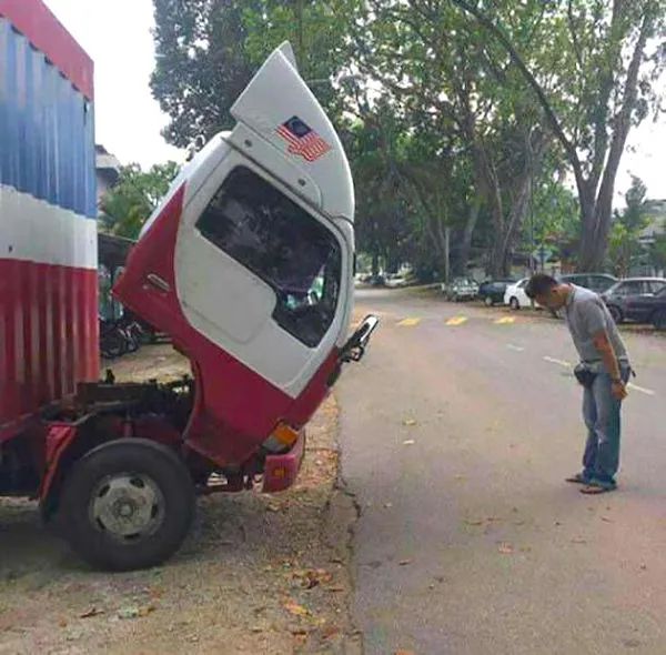 Muž pozoruje sklápanie kabíny kamióna zaparkovaného na kraji cesty pod zelenými stromami.