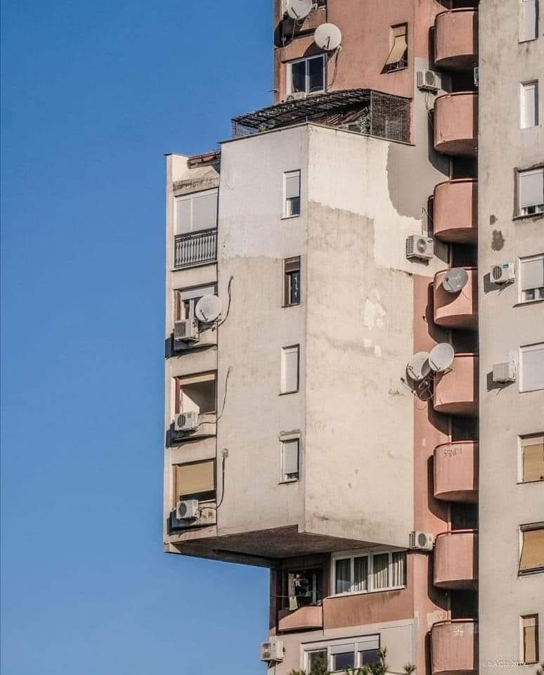 Budova s nevšednou architektúrou a vyčnievajúcim blokom, posiate satelitmi a klimatizáciami, proti modrej oblohe.