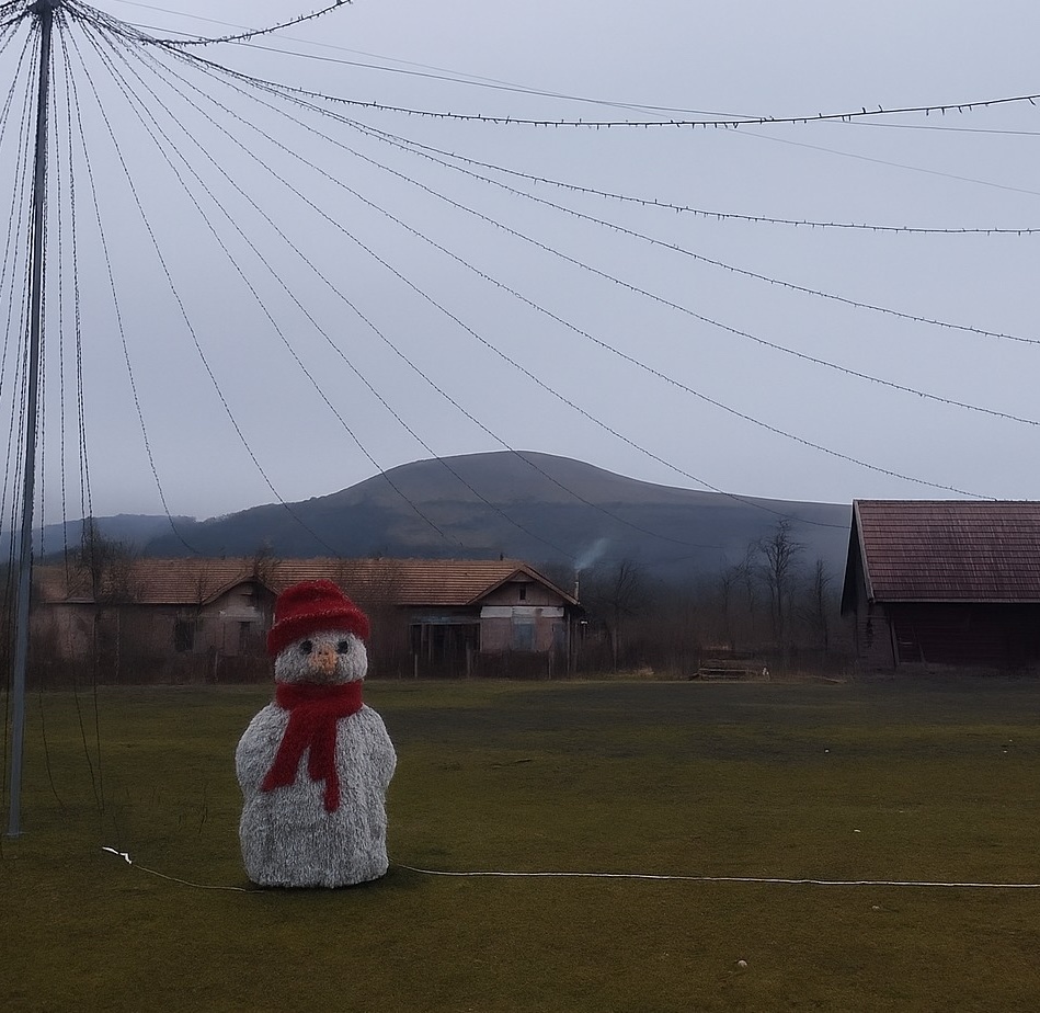 Snehuliak s červeným klobúkom a šálom v zasneženej krajine pri opustených budovách a horách v pozadí.