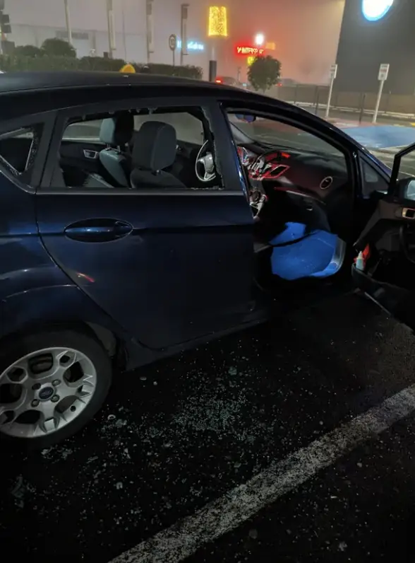 Auto s otvorenými dverami na parkovisku, rozbité sklo na zemi.