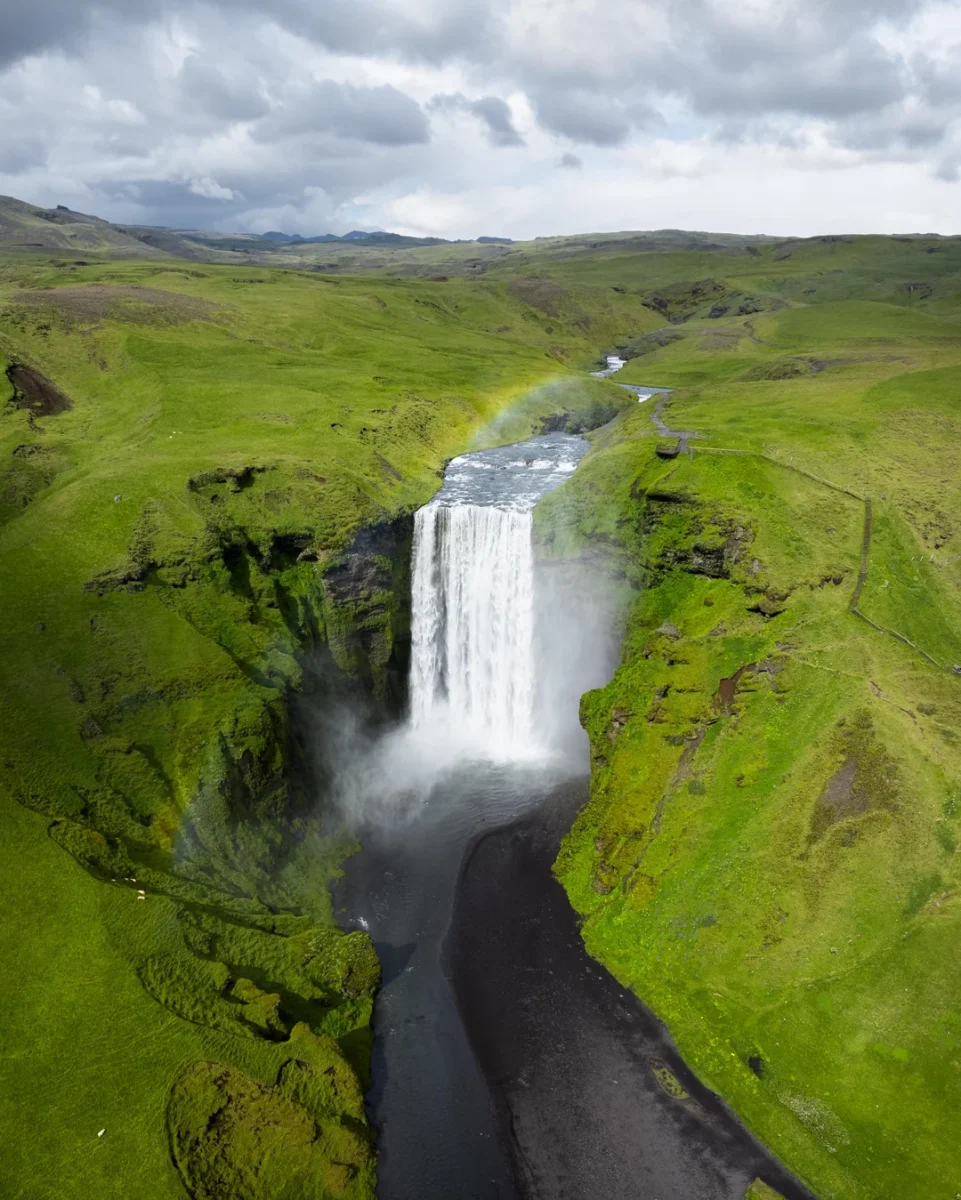 Vodopád obklopený zelenými lúkami a tmavými skalami pod zamračenou oblohou s dúhou, vytvárajúci úchvatný pohľad.