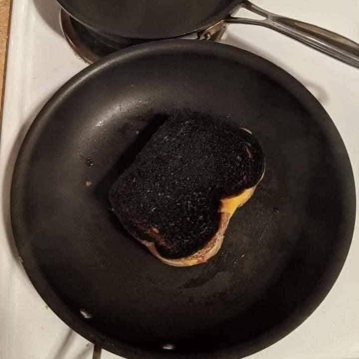 Spálený sendvič na čiernej panvici, syr presakuje na bokoch, dôkaz nešťastného kulinárskeho pokusu.