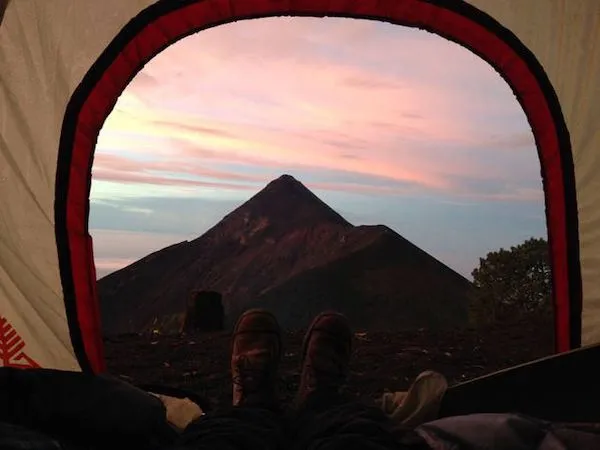 Pohľad na sopku z vnútra stanu pri západe slnka, zatiaľ čo niekto odpočíva nohami vystrčenými smerom k otvoru.