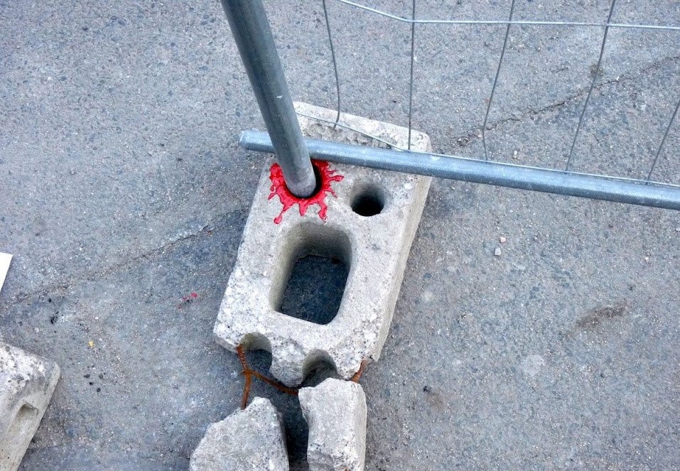 Betónový blok so žrďou, okolo ktorej je červená farba, umiestnený na asfaltovom povrchu ako časť plotu.