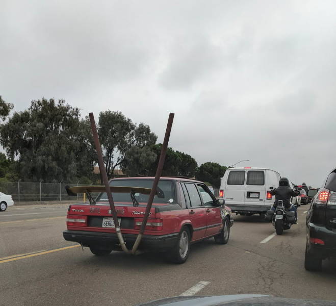Červené auto s vysokými výfukmi na ceste, obklopené ďalšími vozidlami pod zamračenou oblohou.