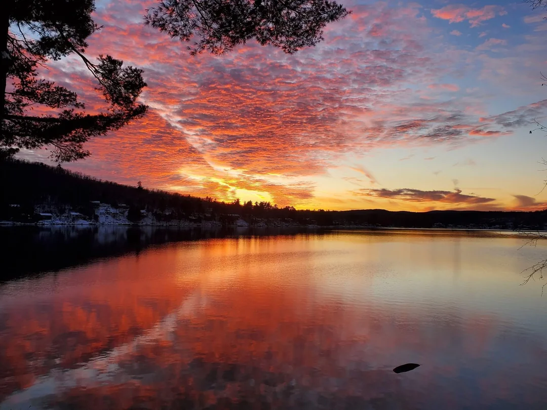 Farebné súmrakové nebo sa odráža na hladine pokojného jazera obklopeného zasneženými kopcami a stromami.