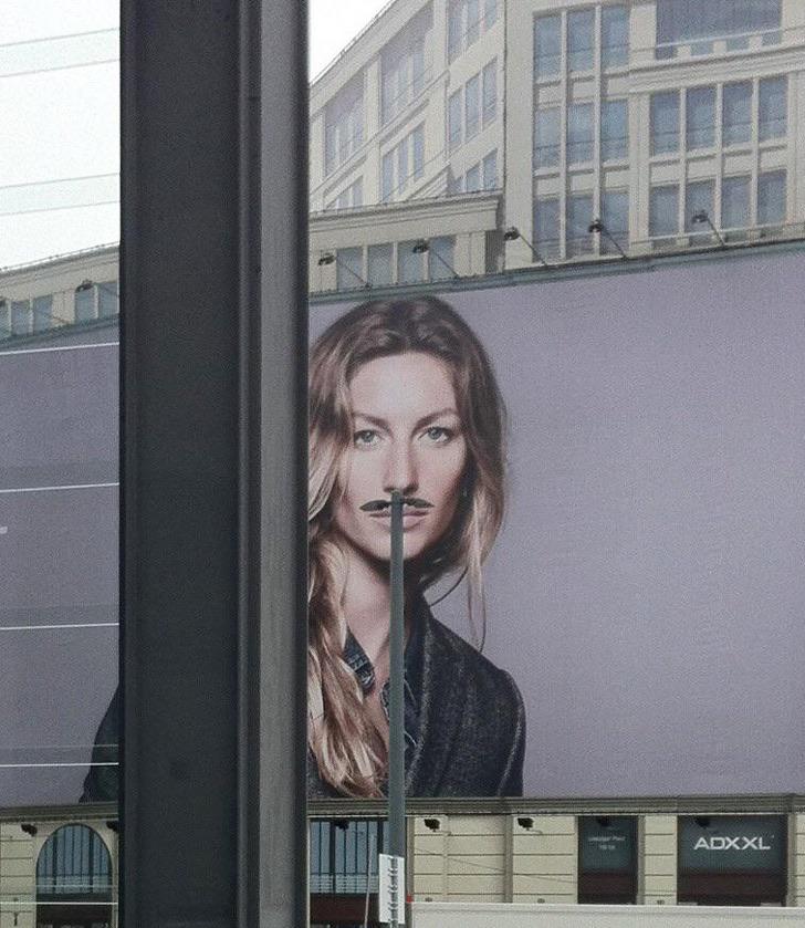 Obrovský billboard ukazuje ženu, na ktorej tvári stĺpik vytvára efekt fúzov pred budovou.