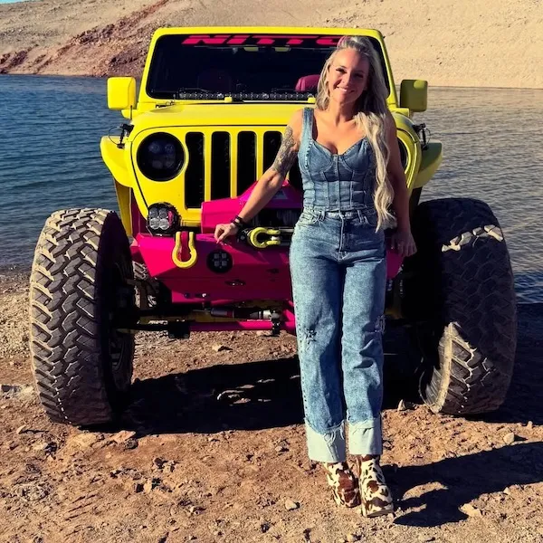 Žena v riflovom odeve stojí pred žltým džípom pri jazere. Letná scéna s terénnym vozidlom a prírodou v pozadí.