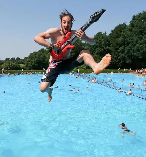 Osoba skáče so skladacou gitarou do bazéna, letná zábava na pláži, modrá voda, slnečný deň.