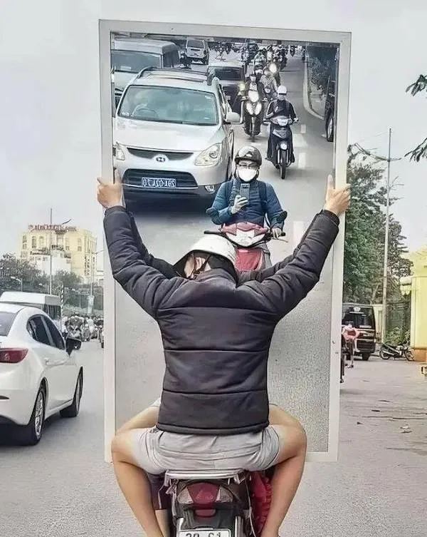 Motorkár prepravuje veľké zrkadlo, v ktorom sa odráža hustá premávka. Kreatívna manipulácia s perspektívou.