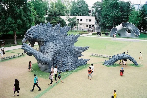 Socha obrieho plaza v parku, okolo ktorej kráčajú návštevníci na zelenom trávniku s modernou architektúrou v pozadí.