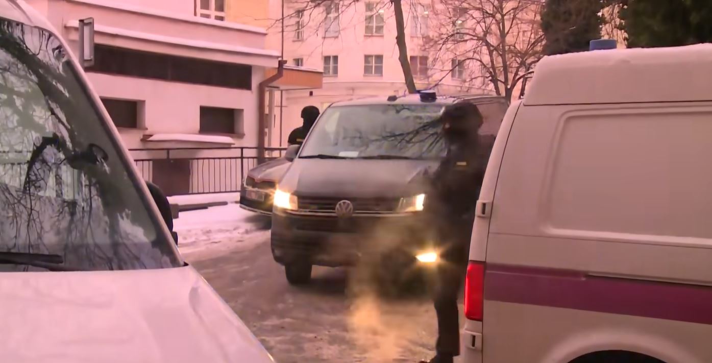 Vozidlá na zasneženej ulici pred budovou za chladného počasia, ľudia v tmavom oblečení, zimná scenéria.