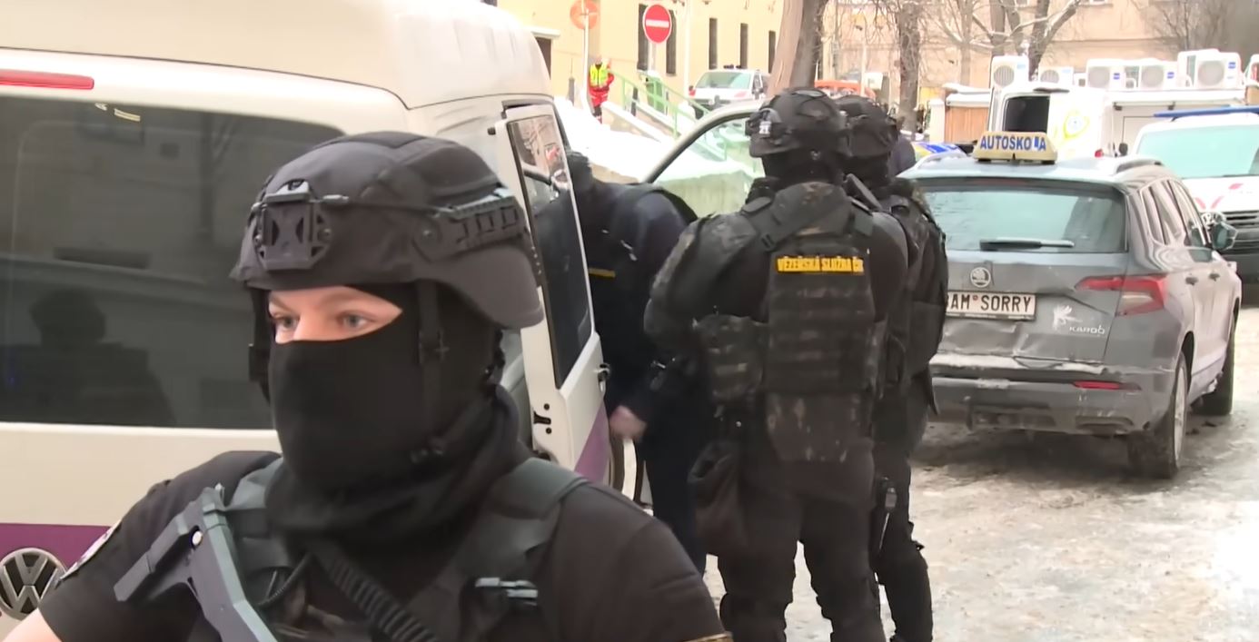 Ozbrojení príslušníci zásahovej jednotky pri zásahu v zimnom prostredí, s dodávkou a autami na pozadí.