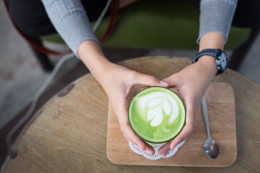 Osoba drží šálku matcha čaju s umením latte na drevenom stolíku, vedľa je lyžička.