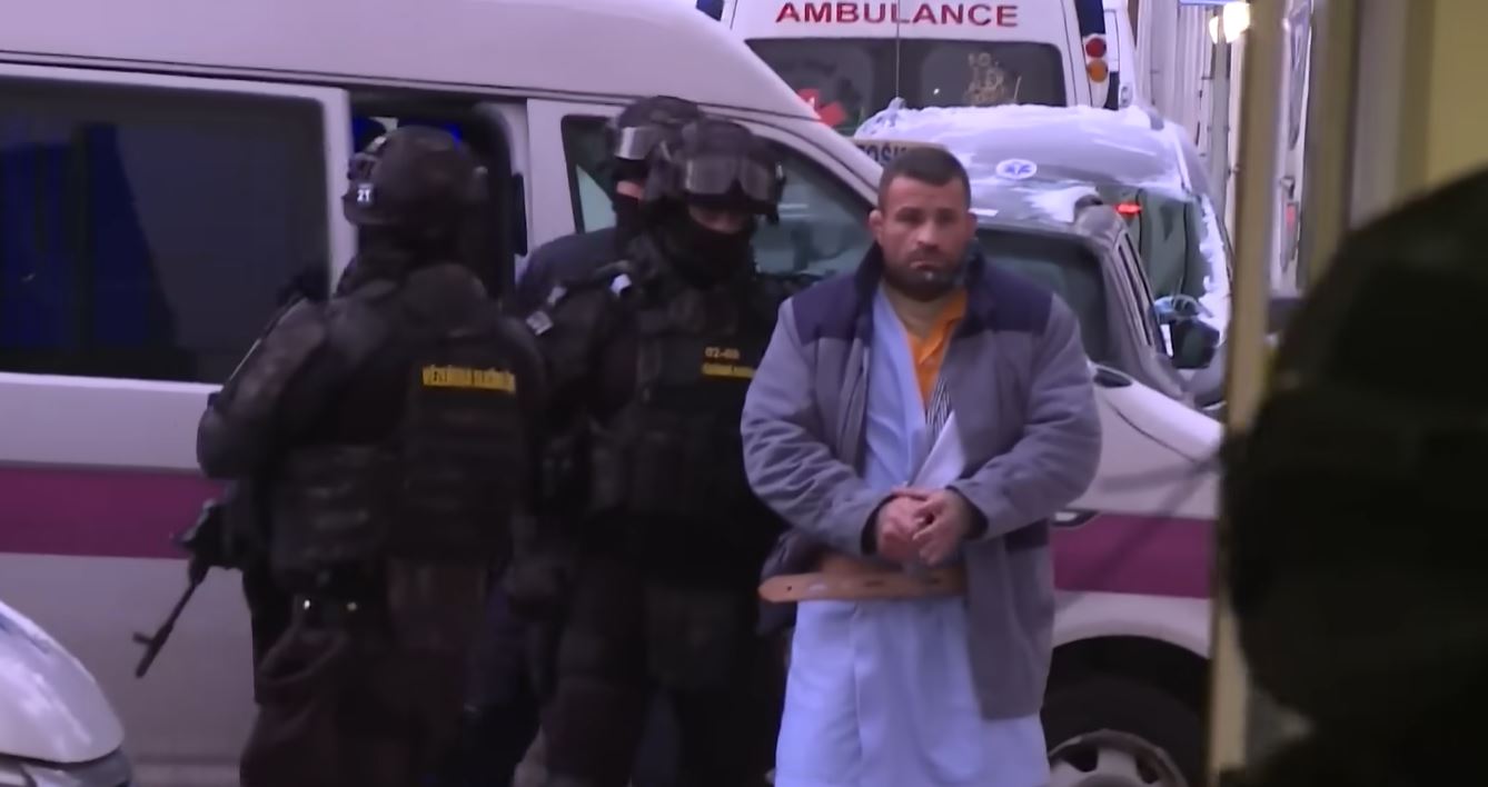 Bezpečnostné zložky vedú muža v putách k sanitke na ulici, doprovod policajtov v maskách a uniformách.