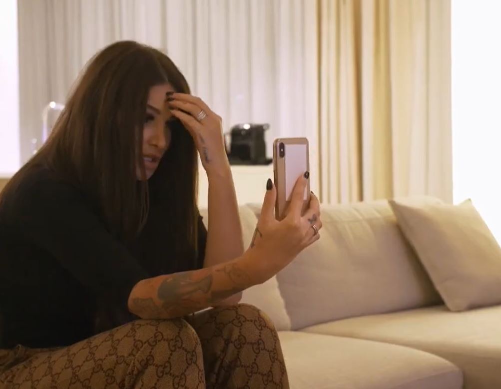 Žena s tetovaniami sedí na pohovke a telefonuje cez smartphone, vyzerá znepokojene.