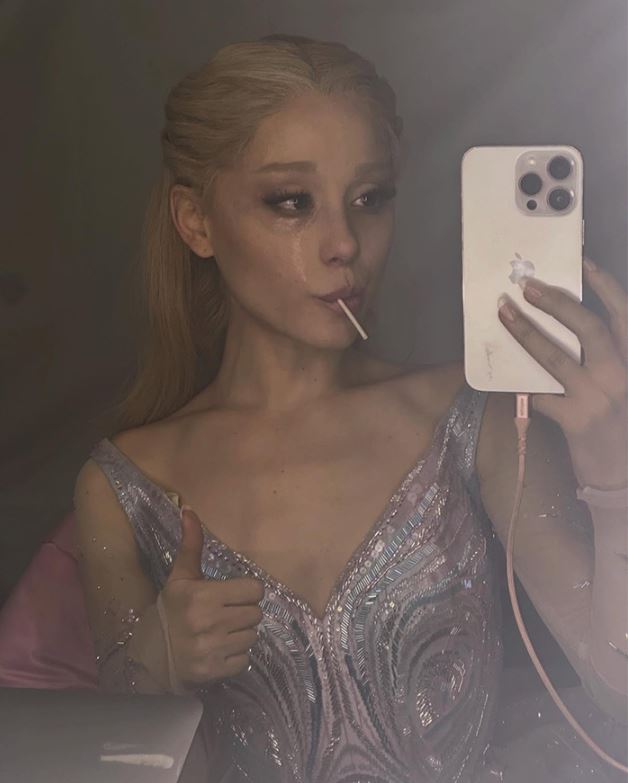 Osoba v elegantných šatách robí selfie s palcom hore a lízatkom, vyjadruje spokojnosť v šatni počas večerného podujatia.