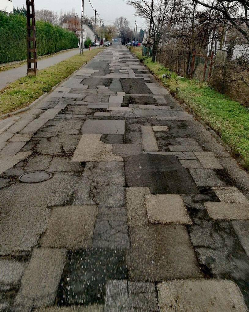 Nerovnomerná cesta s množstvom záplat a prasklín vedúca cez malebnú obytnú oblasť.