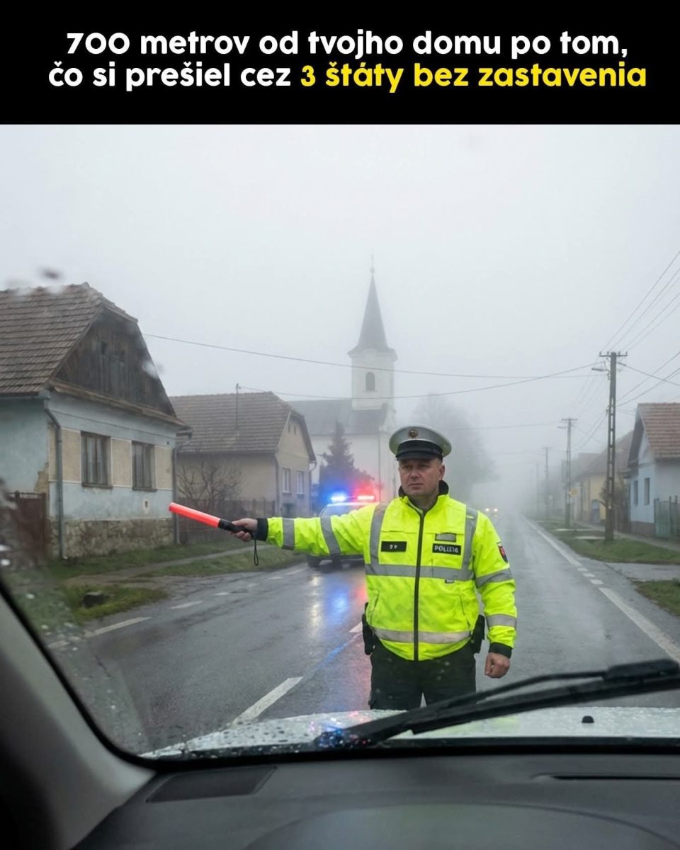 Policajt zastavuje auto na dedinskej ceste v hmle, v pozadí vidno kostol.