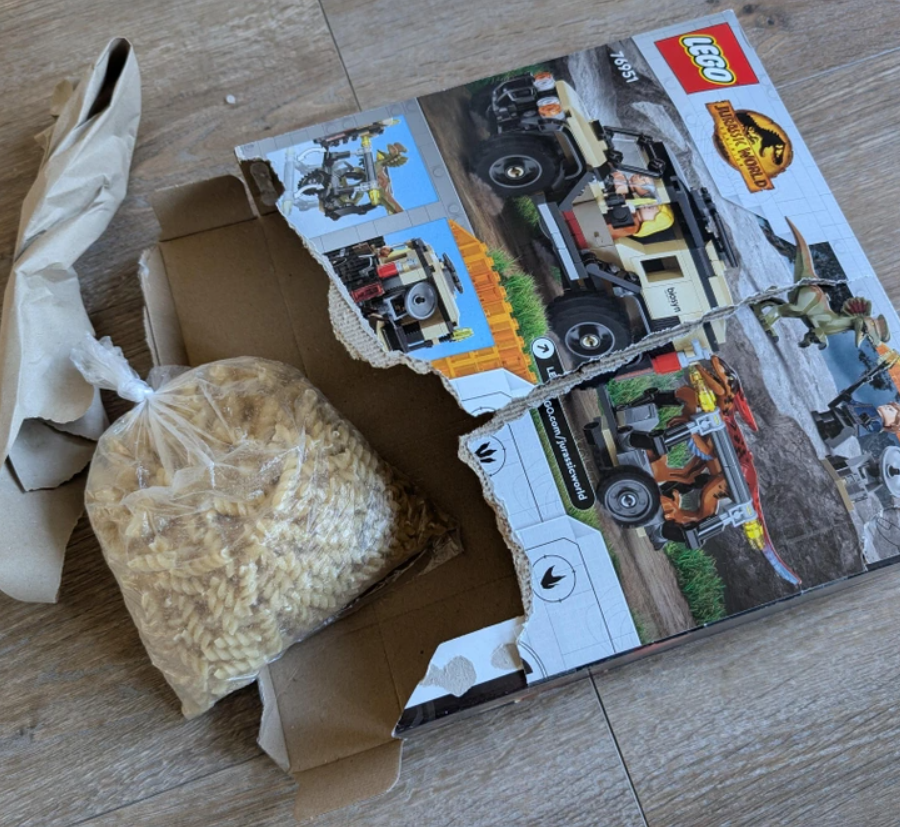 Roztrhnutá LEGO krabica so setom Jurassic World a vedľa igelitové vrecko plné sušených cestovín.