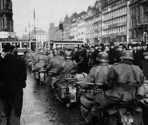 Ozbrojení vojaci na motocykloch prechádzajú mestskými ulicami plnými ľudí, historická čiernobiela fotografia.