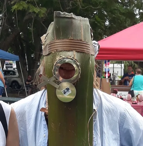 Osoba s maskou z prírodného materiálu na outdoorovom trhu, ľudia v pozadí pod stánkami.