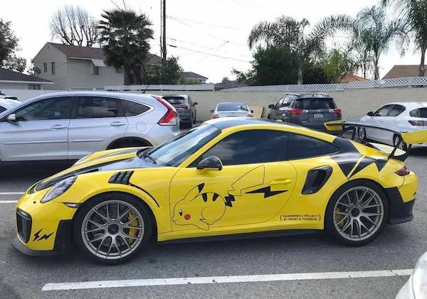 Žlté auto Porsche s motívom Pikachu na parkovisku obklopené inými autami.