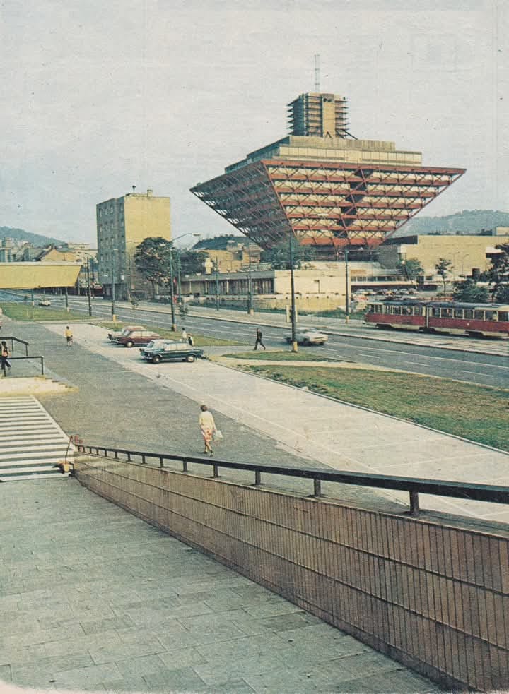 Ľudia a autá pred obrátenou pyramídou, ikonickou budovou v Bratislave, v slnečný deň.