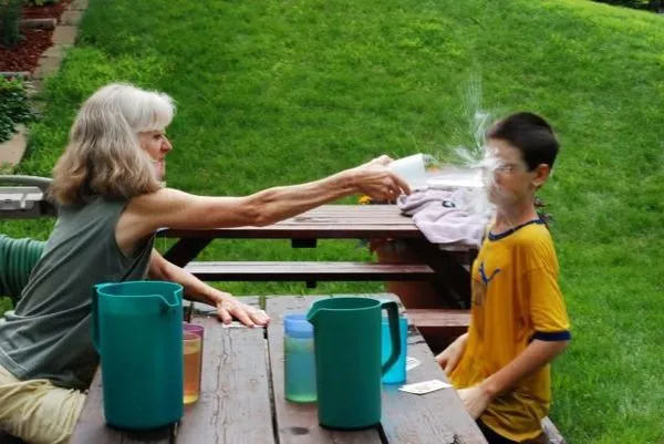 Žena s úsmevom strieka vodu na chlapca sediaceho pri piknikovom stole vonku na tráve.