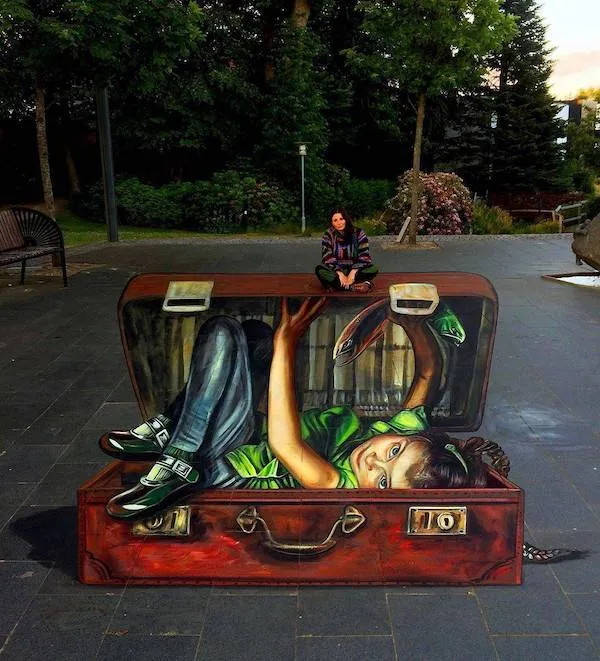3D ilúzia dieťaťa v kufri na ulici, na vrchu sediaca žena. Street art vo verejnom priestore.