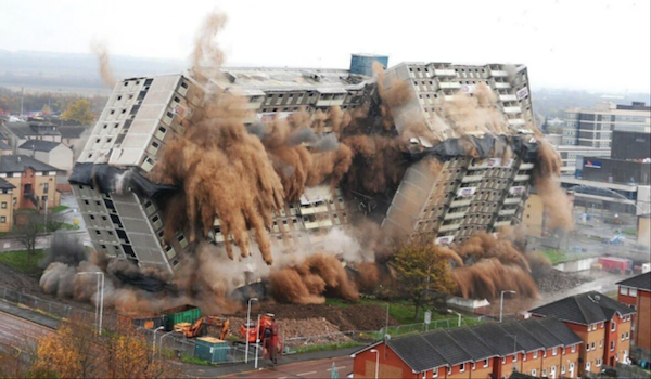 Budova počas kontrolovanej demolácie, padá obklopená prachom, s okolitými stavbami v pozadí.