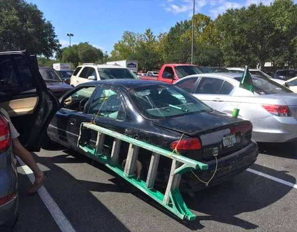 Auto s rebríkom pripevneným na bok stojí na parkovisku, kreatívne riešenie prepravy.