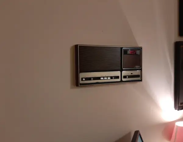 Starožitný nástenný rádio prijímač s analógovými ovládačmi a integrovanými hodinami na bielej stene, osvetlený lampou.