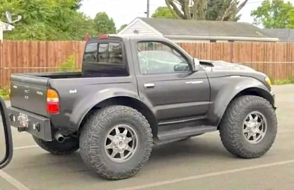 Modifikované pick-up vozidlo s veľkými terénnymi pneumatikami na parkovisku pred dreveným plotom.