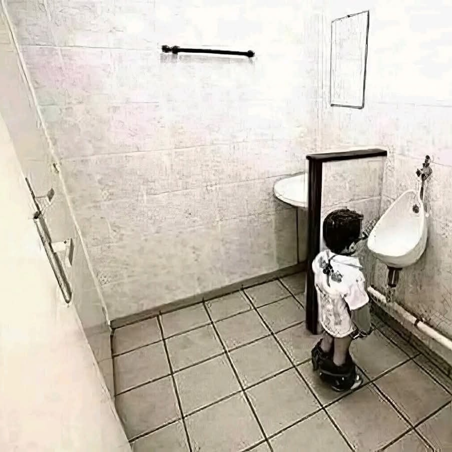 Malý chlapec stojí pred zrkadlom v kúpeľni pri pisoári v zábavnej optickej ilúzii.