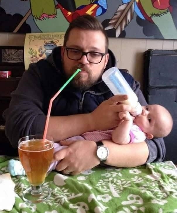 Muž sedí pri stole, pije nápoj zo pohára a kŕmi bábätko fľaškou mlieka.